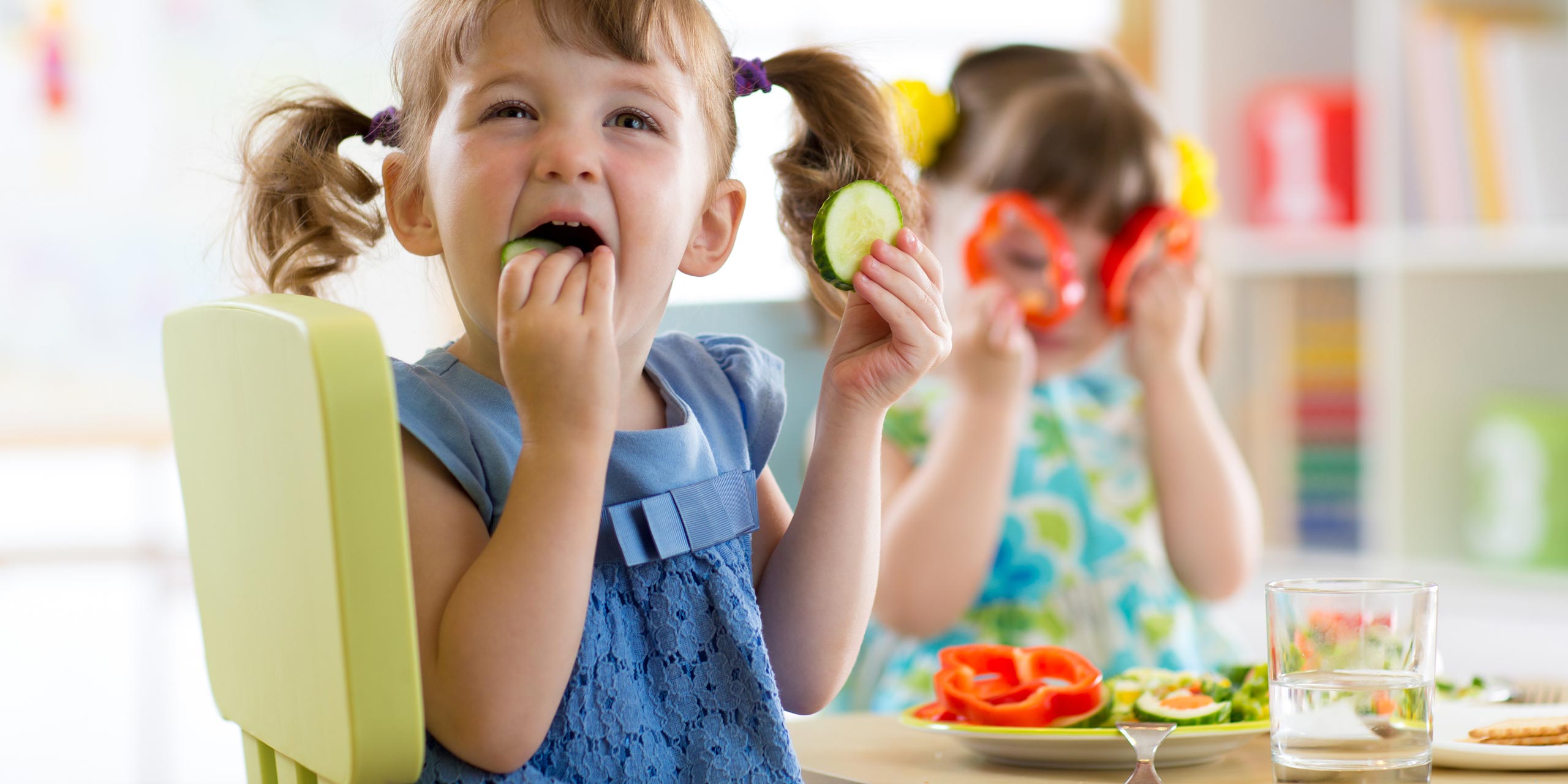 Zwei kleine Mädchen am Esstisch. Das Kind im Vordergrund isst Gurke, das Kind im Hintergrund benutzt zwei Paprikascheiben als Spielzeugbrille.