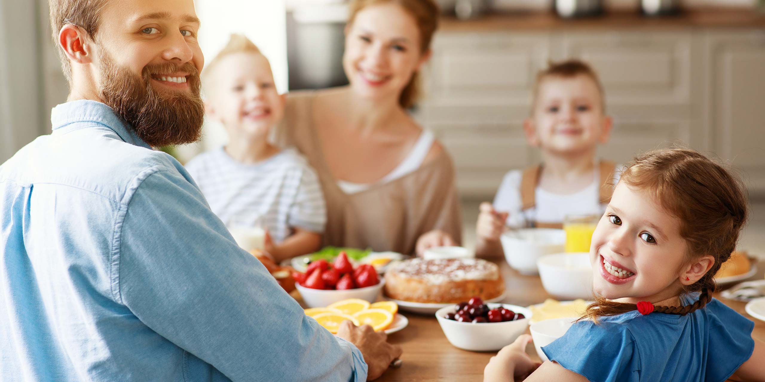 Junge Familie am Tisch mit verschiedenen Backwaren und Obst, alle lächeln in die Kamera.